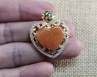 Puede incluir: Un colgante de corazón de oro con una piedra preciosa de corazón de color naranja claro en el centro. El colgante está rodeado por un diseño de filigrana decorativo con pequeñas piedras transparentes.