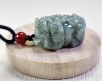 Puede incluir: Un colgante de jade verde tallado en forma de criatura mítica, con detalles intrincados. El colgante está unido a un cordón negro con una cuenta roja.