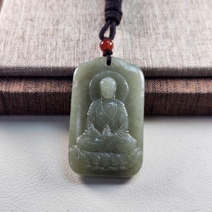 Puede incluir: Un colgante rectangular de jade verde claro con la figura tallada de un Buda sentado sobre una base de loto. El colgante cuelga de un cordón marrón oscuro con una pequeña cuenta roja pulida situada directamente sobre la piedra tallada.