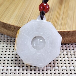 May include: White jade pendant with a circular opening in the center and eight sides. The pendant has Chinese characters carved into the surface. The pendant is attached to a black cord with a red bead.