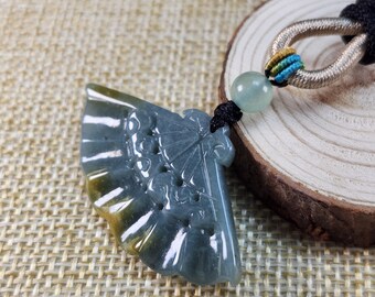 Puede incluir: Un colgante de jade tallado en forma de abanico con un esquema de color verde y amarillo. El colgante está unido a un cordón negro con una cuenta verde.