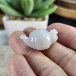 Könnte beinhalten: Ein kleiner, weißer Jade-Teekannen-Anhänger. Die Teekanne hat einen abgerundeten Körper mit Ausguss und Henkel. Der Jade ist durchscheinend, mit einer glatten, polierten Oberfläche. Die Teekanne wird in einer Hand gehalten.