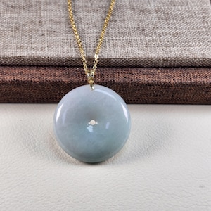 May include: A round, pale green jade pendant with a small central hole, suspended from a delicate gold chain. The jade has a smooth, polished surface and a subtle color gradient. The necklace is displayed on a neutral background.