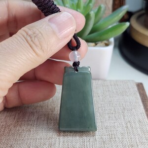 Puede incluir: Colgante de jade verde oscuro con forma rectangular y parte superior decorativa. El colgante está suspendido de un cordón negro con una pequeña cuenta rosa claro. El jade tiene una superficie lisa y pulida.
