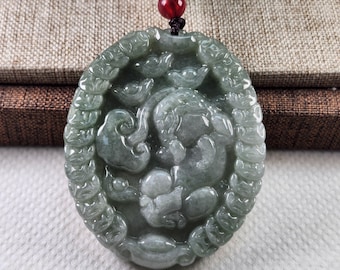 Puede incluir: Colgante ovalado de jade con un diseño tallado. El colgante presenta una figura central rodeada de formas de nubes y un borde adornado. El jade es de color verde apagado, y el colgante está suspendido de un cordón negro con una cuenta roja.