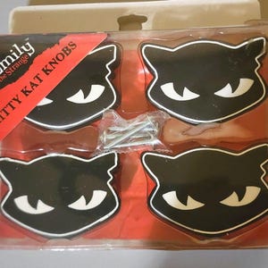 May include: Four black and white cat-shaped cabinet knobs in a clear plastic package. The knobs have a black cat face design with white eyes and a white outline. The package includes screws and the text "Emily the Strange Kitty Kat Knobs".