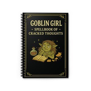 Puede incluir: Un cuaderno con espiral y cubierta negra con el texto "GOBLIN GIRL + SPELLBOOK OF + CRACKED THOUGHTS" en letras doradas. La ilustración de la portada representa un duende leyendo un libro, con una botella de poción y galletas. El cuaderno tiene un fondo blanco.