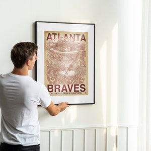 May include: A person hangs a framed poster on a white wall. The poster is a vintage-style illustration of a baseball stadium with the text "Atlanta Braves 2021 World Series Champions".