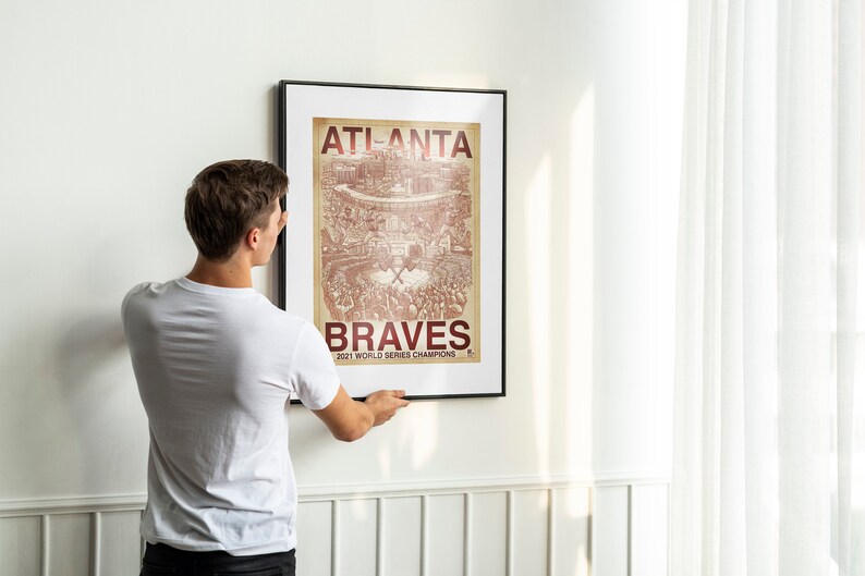 May include: A person hangs a framed poster on a white wall. The poster is a vintage-style illustration of a baseball stadium with the text "Atlanta Braves 2021 World Series Champions".