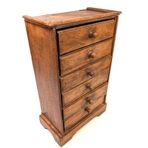 May include: A small, antique wooden cabinet with six drawers. The cabinet is made of a light brown wood and has a simple, classic design. Each drawer has a small, round wooden knob. The cabinet is a vintage piece.