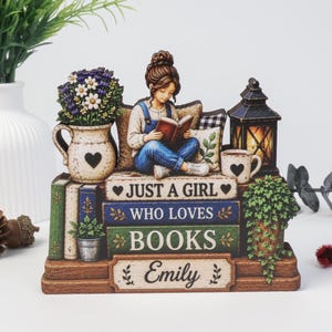 Könnte beinhalten: Eine dekorative Tischfigur, die ein Mädchen zeigt, das ein Buch liest, während es auf einem Bücherstapel sitzt. Die Bücher tragen die Aufschrift JUST A GIRL WHO LOVES BOOKS und den Namen Emily. Enthält eine Laterne, eine Blumenvase und eine Tasse.