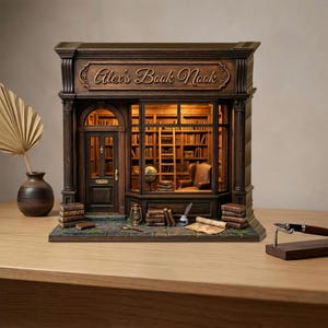 May include: A miniature, detailed model of a bookstore, featuring the text "Alex's Book Nook" above the window. The interior is lit, showcasing bookshelves, a ladder, and a reading chair. Books and accessories are arranged on the exterior.