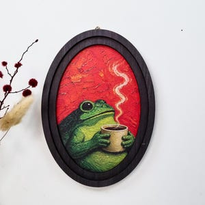 May include: An oval-framed oil painting featuring a green frog holding a beige mug with rising steam. The background is a textured, vibrant red. The frame is dark wood, and the artwork displays visible brushstrokes throughout the composition.