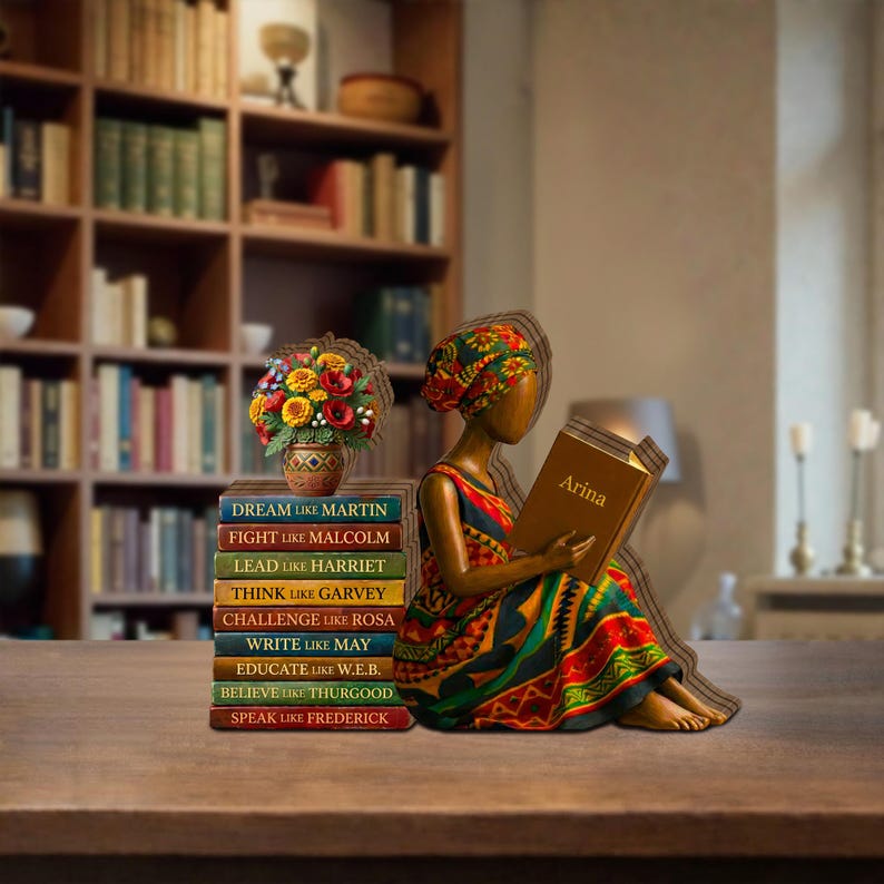 May include: A wooden figurine of a woman reading a book with the name "Arina" on the cover. Beside her is a stack of books with inspirational phrases like "Dream like Martin" and a vase of flowers. The woman wears a colorful dress and head wrap.