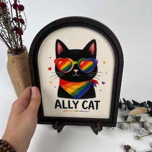 Op de afbeelding: Een illustratie van een zwarte kat in een boogvormige lijst van donker hout. De kat draagt hartvormige regenboogzonnebril en een regenboogbandana. De tekst ALLY CAT staat in zwarte letters onder de kat, vergezeld door kleine hartjes en sterren.