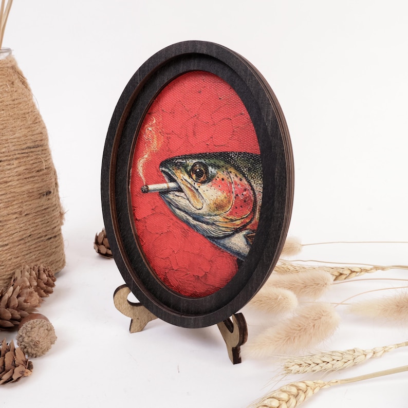 May include: An oval-shaped dark wood frame containing a painting of a trout fish with a cigarette in its mouth. The fish is set against a textured red background. The frame is supported by a small, matching dark wood stand on a flat surface.