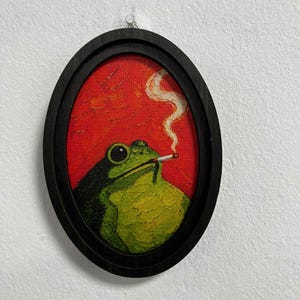 May include: An oval-shaped framed painting featuring a green frog with large black eyes holding a lit cigarette in its mouth. The frog is set against a vibrant red background with a visible trail of white smoke rising from the cigarette.