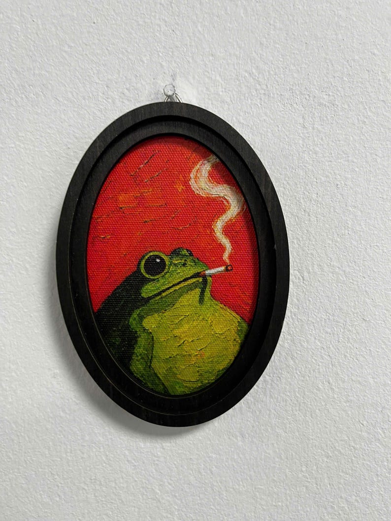 May include: An oval-shaped framed painting featuring a green frog with large black eyes holding a lit cigarette in its mouth. The frog is set against a vibrant red background with a visible trail of white smoke rising from the cigarette.