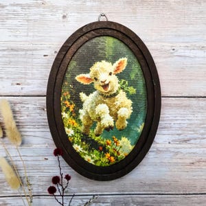 Puede incluir: Un adorno de pared ovalado que presenta una ilustración impresa de un cordero blanco saltando a través de un campo de hierba verde y flores amarillas. La obra está enmarcada en un marco de madera marrón oscuro con un gancho metálico arriba.