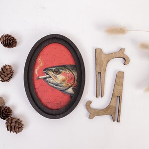 May include: An oval-shaped dark wood frame containing a print of a fish with a cigarette in its mouth against a textured red background. Two separate wooden display stands are positioned to the right of the frame, and pinecones are scattered.