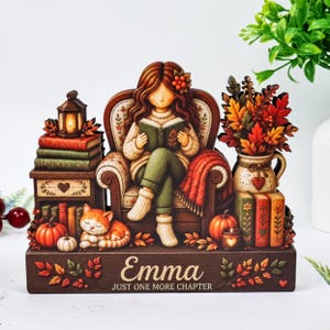 Puede incluir: Un letrero decorativo de madera para mesa que muestra a una niña leyendo un libro en un sillón, un gato naranja dormido, pilas de libros, calabazas y un jarrón con hojas de otoño. La base tiene grabado el nombre Emma y el texto JUST ONE MORE CHAPTER.