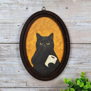 May include: An oval-framed painting featuring a black cat with yellow eyes against a textured mustard yellow background. The cat is holding a white roll of toilet paper in its front paws. The frame is dark brown wood with a metal hanging hook.