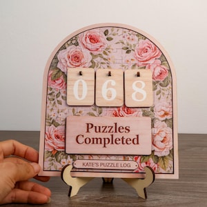 May include: An arched wooden puzzle tracker featuring a floral rose pattern with three light wood tiles displaying the number 068. A rectangular plaque reads Puzzles Completed, and a smaller label below reads KATE'S PUZZLE LOG. The item rests on a stand.