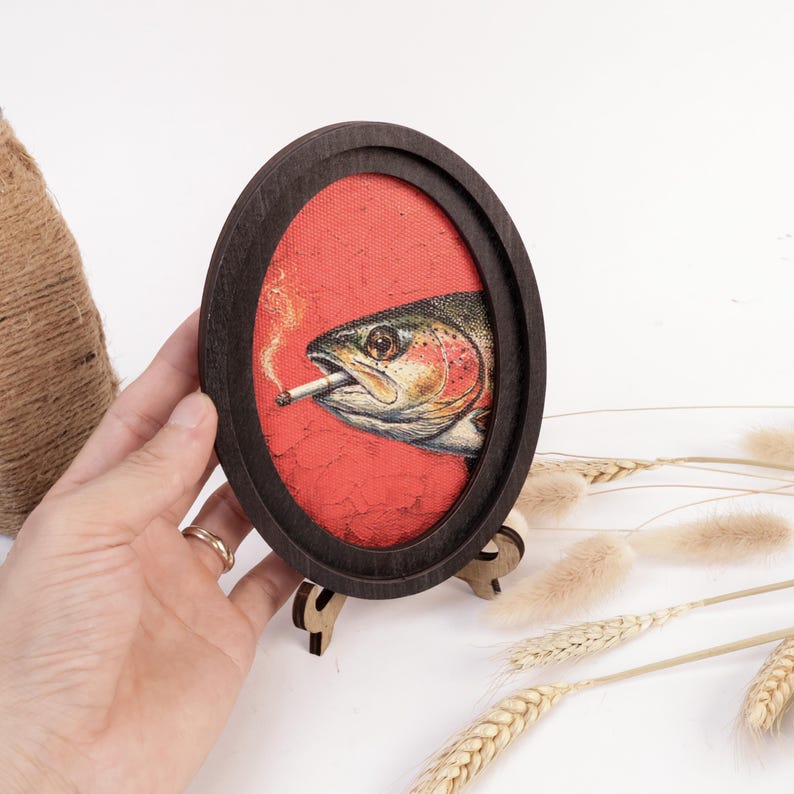 May include: An oval-shaped dark wood frame containing a canvas print of a fish with a cigarette in its mouth. The fish is depicted in profile against a textured red background, with a small wisp of smoke rising from the lit cigarette tip.