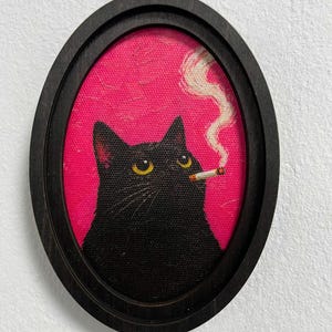 Puede incluir: Una pieza de arte mural con marco ovalado que muestra un gato negro de ojos amarillos con un cigarrillo en la boca. El gato está sobre un fondo rosa intenso, con una estela de humo blanco que se eleva desde el cigarrillo hacia el aire.