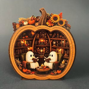 May include: A pumpkin-shaped wooden diorama featuring two white ghost figures sitting in armchairs and reading books. The interior depicts a library with shelves of books, warm lighting, and autumn decorations including sunflowers and orange pumpkins.