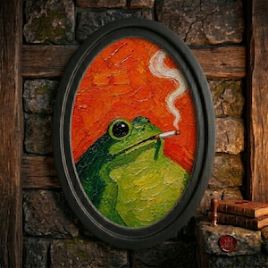 May include: An oval-framed oil painting featuring a green frog with a cigarette in its mouth, exhaling a trail of white smoke against a textured, vibrant orange background. The artwork is encased in a simple black frame with visible brushstrokes.