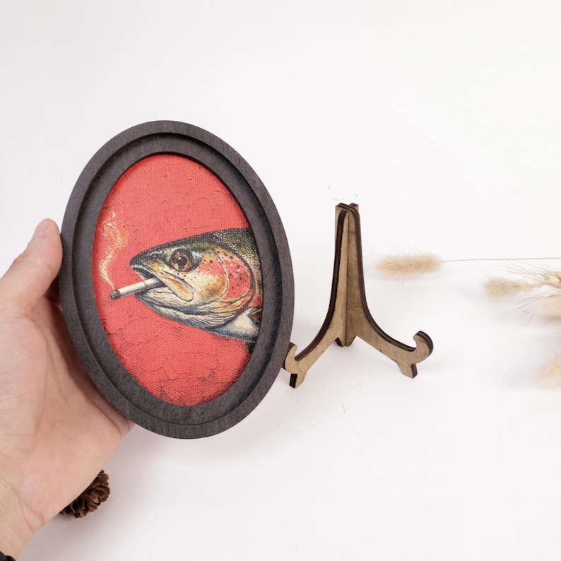 May include: An oval-shaped dark wood frame containing a canvas print of a fish with a cigarette in its mouth. The background behind the fish is a textured red color. The frame is displayed with a matching dark wood easel stand for support.