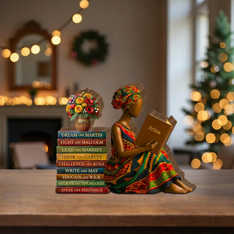 May include: A wooden figurine of a person reading a book titled "Arina", wearing a colorful dress and head wrap. Beside the figure is a stack of books with titles like "Dream Like Martin" and a vase of flowers. The scene is set on a wooden table.