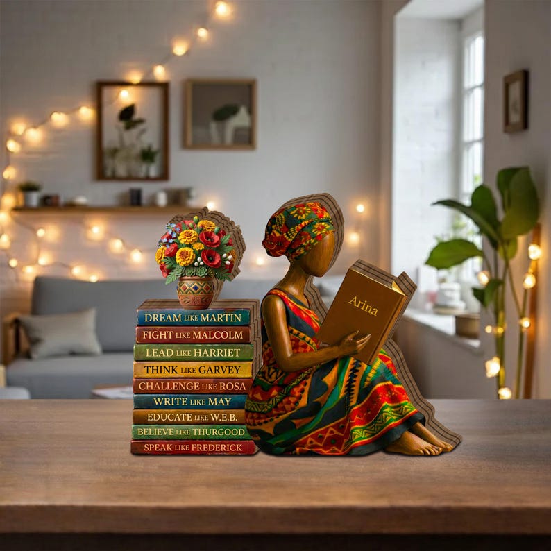 May include: A wooden figurine of a person reading a book with the name "Arina" on the cover. The figurine is wearing a colorful dress and head wrap. A stack of books with inspirational quotes and a vase of flowers are next to the figurine.