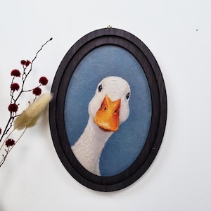 May include: An oval-framed oil painting featuring a white goose with a bright orange beak set against a textured blue background. The goose is positioned centrally, looking forward, and is enclosed in a dark, smooth wooden frame.