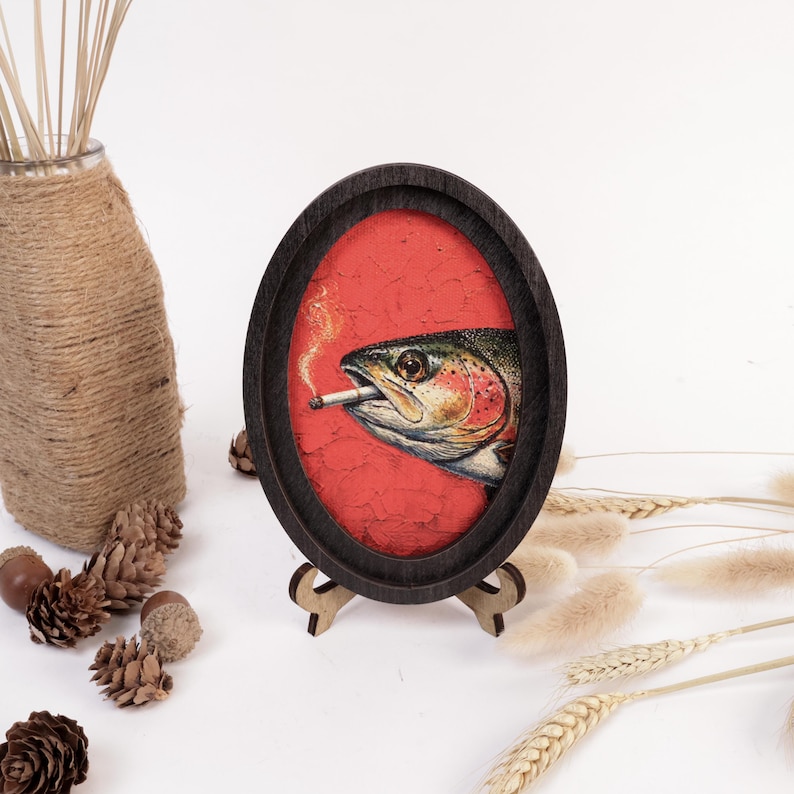 May include: An oval-shaped dark wooden frame containing a detailed illustration of a trout fish with a cigarette in its mouth, set against a textured red background. The frame is supported by a small wooden stand on a white surface.