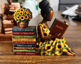 May include: A figurine of a woman in a yellow sunflower dress reading a book with the name "Jackie" on the cover. A stack of books with inspirational phrases and a vase of sunflowers are nearby. Another figurine of a woman is seated in a chair.