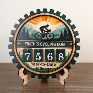 Könnte beinhalten: Ein kreisförmiges, zahnradförmiges Radsport-Logbuch aus Holz mit der Silhouette eines Radfahrers vor einem Bergsonnenaufgang. Der Text lautet ERICH'S CYCLING LOG und Year-to-Date Mileage. Vier dunkelgrüne Klappkarten zeigen die Zahl 7568.