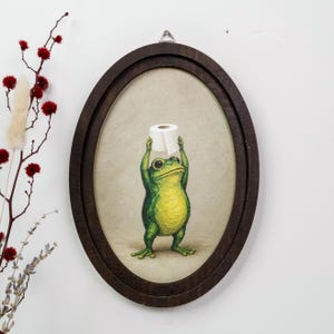 Puede incluir: Una pieza de arte mural de forma ovalada que presenta una pintura de una rana verde de pie, sosteniendo un rollo de papel higiénico blanco sobre su cabeza. La obra está enmarcada en un marco de madera marrón oscuro con un gancho de metal.