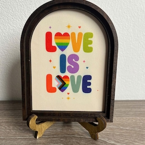 Puede incluir: Un marco de madera arqueado que contiene una impresión en lienzo con el texto LOVE IS LOVE. Las letras son coloridas, con la O del primer LOVE como un corazón arcoíris y la O del segundo LOVE como un corazón progress pride. Pequeñas estrellas y corazones rodean el texto.