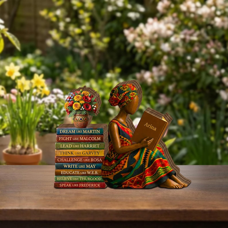 May include: A wooden figurine of a person reading a book titled "Arina", wearing a colorful dress and head wrap. A stack of books with inspirational phrases like "Dream like Martin" and a vase of flowers are nearby. The background is a blurred garden.
