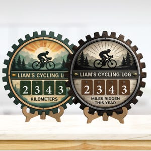 Könnte beinhalten: Zwei hölzerne Fahrradprotokolle in Zahnradform, die "LIAM'S CYCLING LOG" und die Zahl "2343" anzeigen. Eines zeigt Kilometer, das andere gefahrene Meilen. Beide zeigen eine Radfahrer-Silhouette und eine Berglandschaft.