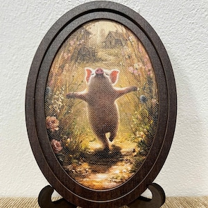 May include: An oval-shaped dark brown wooden frame holds a canvas print of a small pig standing on its hind legs with arms outstretched in a grassy field with flowers and a distant house, supported by a matching dark brown wooden display stand.