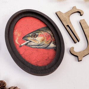 May include: An oval-shaped dark wood frame containing a textured red background with an illustration of a fish smoking a cigarette. Beside the frame is a separate, detached wooden stand piece with a curved, hook-like design.