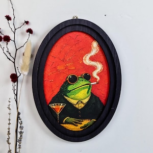 May include: An oval-framed painting featuring a green frog wearing dark round sunglasses and a black suit. The frog is smoking a cigarette with visible smoke rising against a textured red background, while holding a cocktail glass on a table.