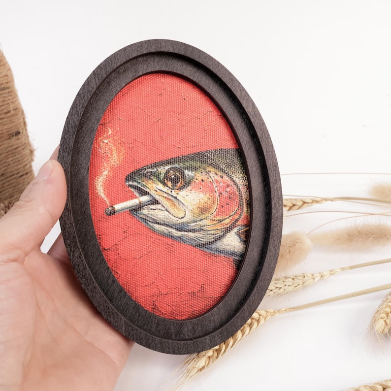 May include: An oval-shaped dark wooden frame containing a canvas print of a fish with a pinkish side and a dark back. The fish is depicted with a lit cigarette in its mouth, with a thin trail of smoke rising against a solid red background.