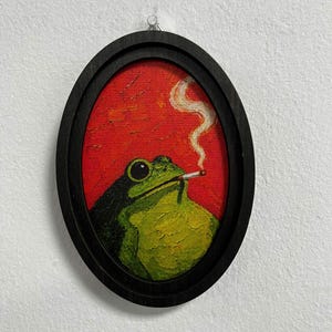 Pode incluir: Uma pintura em moldura oval apresentando um sapo verde com dorso escuro contra um fundo vermelho vibrante. O sapo é retratado com um cigarro na boca, com um fino rastro de fumaça branca subindo em direção ao espaço vermelho texturizado.