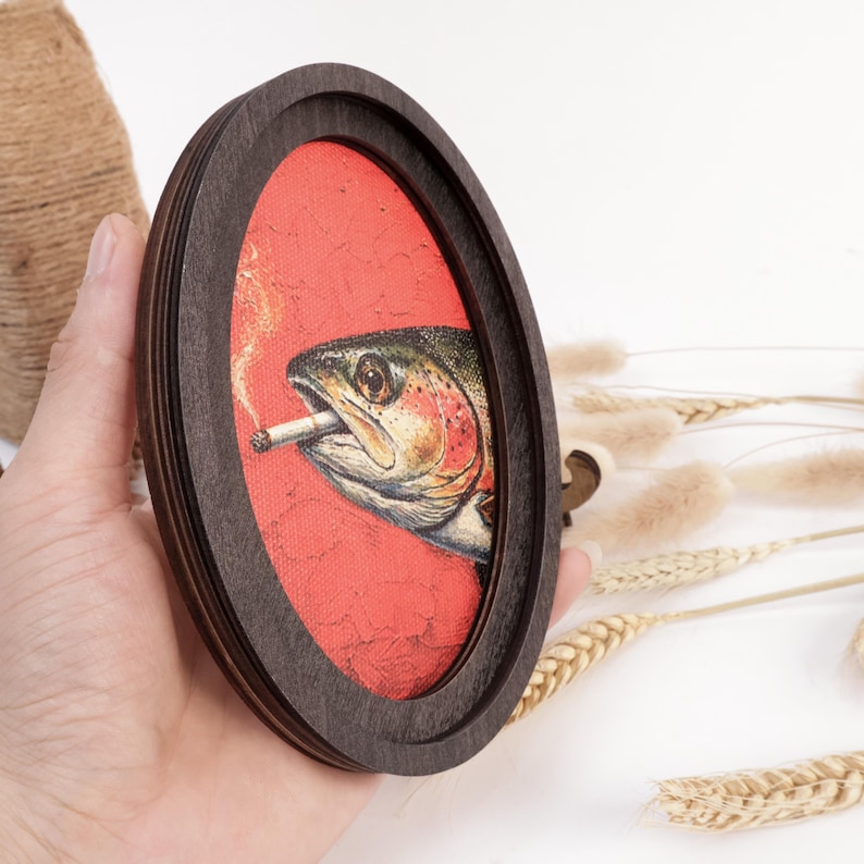 May include: An oval-shaped dark wood frame containing a canvas print of a fish with a cigarette in its mouth. The fish is depicted in profile against a textured, vibrant red background, with a thin wisp of smoke rising from the cigarette.