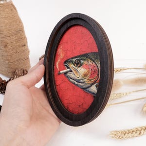 May include: An oval-shaped dark wooden frame containing a canvas print of a fish with a cigarette in its mouth. The fish is depicted in shades of green, silver, and pink against a textured, vibrant red background with a thin trail of smoke.