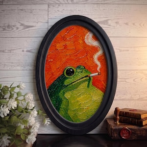 May include: An oval-framed oil painting featuring a green frog with a textured impasto finish. The frog is depicted with a cigarette in its mouth, exhaling a thin trail of white smoke against a vibrant, thick-brushed orange background.
